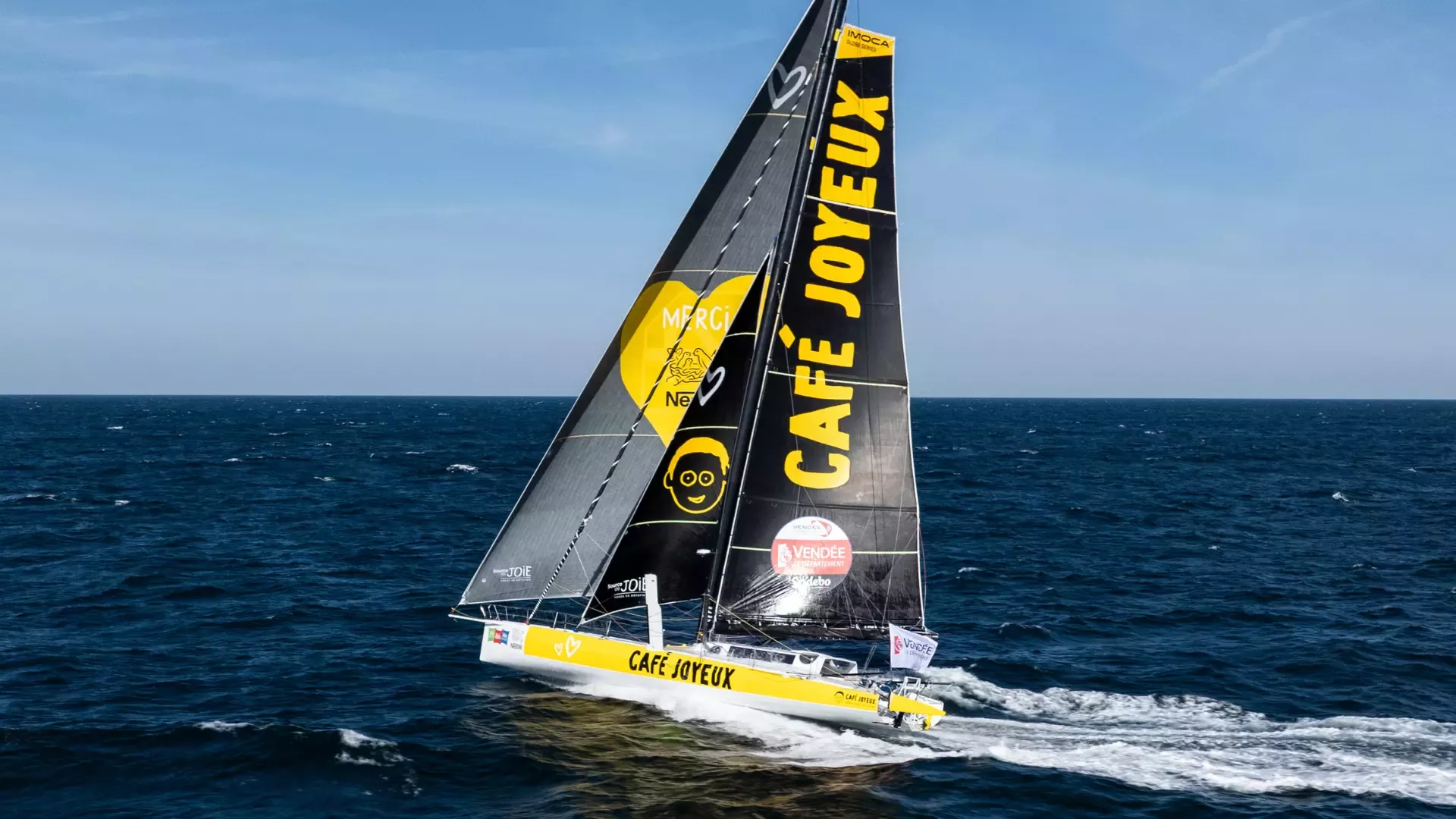Voilier de course au large (IMOCA) noir et jaune « Café Joyeux » naviguant à pleine vitesse sur un océan agité